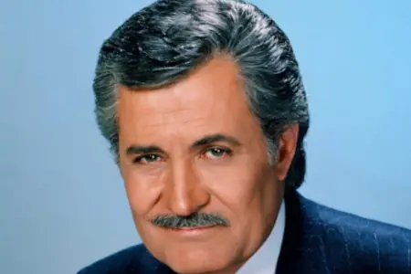 A mature man with well-styled gray hair and a trim mustache looks at the viewer with brown eyes and a slight smile. He sits in a dark blue suit against a light blue background.